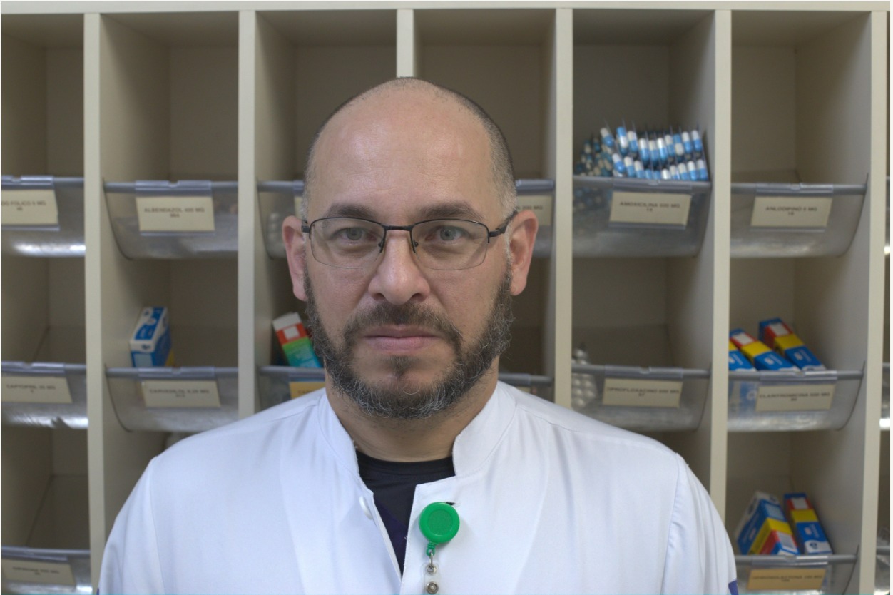 Na imagem, o farmacêutico, um homem careca, olhos claros, de barba e com óculos, usa jaleco branco e atrás dele há um armário com medicamentos